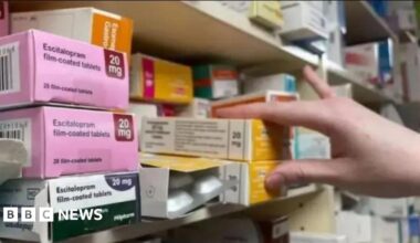Boxes of medication on a shelf in a chemist, with a hand reaching for two boxes.