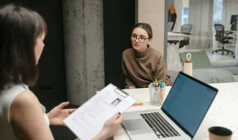 Getty Images Woman holds CV of a young woman sat in front of her in an office setting