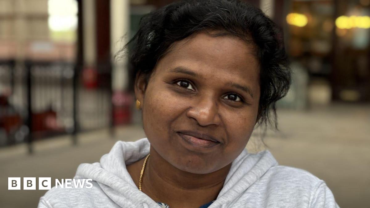 Janet is wearing a light grey jumper and has gold earrings and chain.