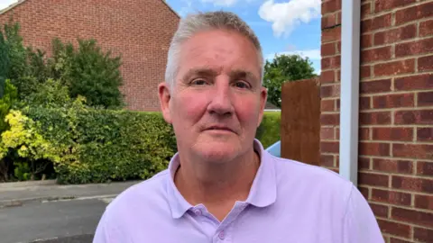 BBC Mark Hemphill stood on a residential street. He is wearing a purple polo tshirt and is staring into the camera.