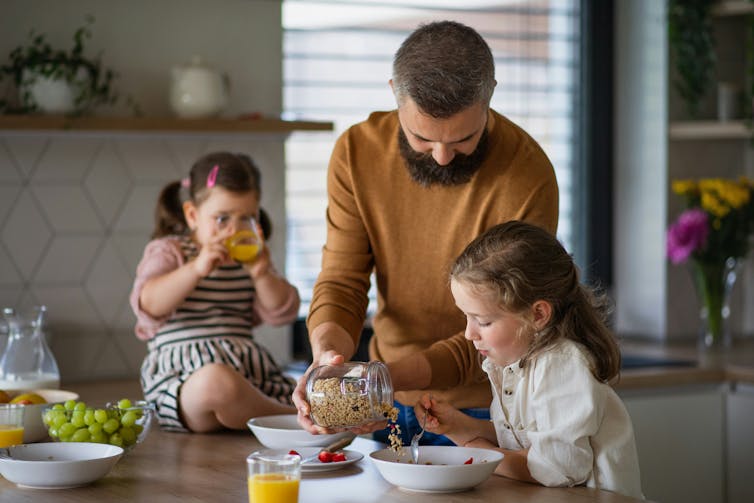 A man in a kitchen with two little girls, pouring cereal into a bowl for one of them