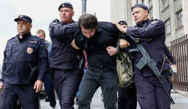 Three policemen in uniform lead away a bearded man in jeans and a black shirt