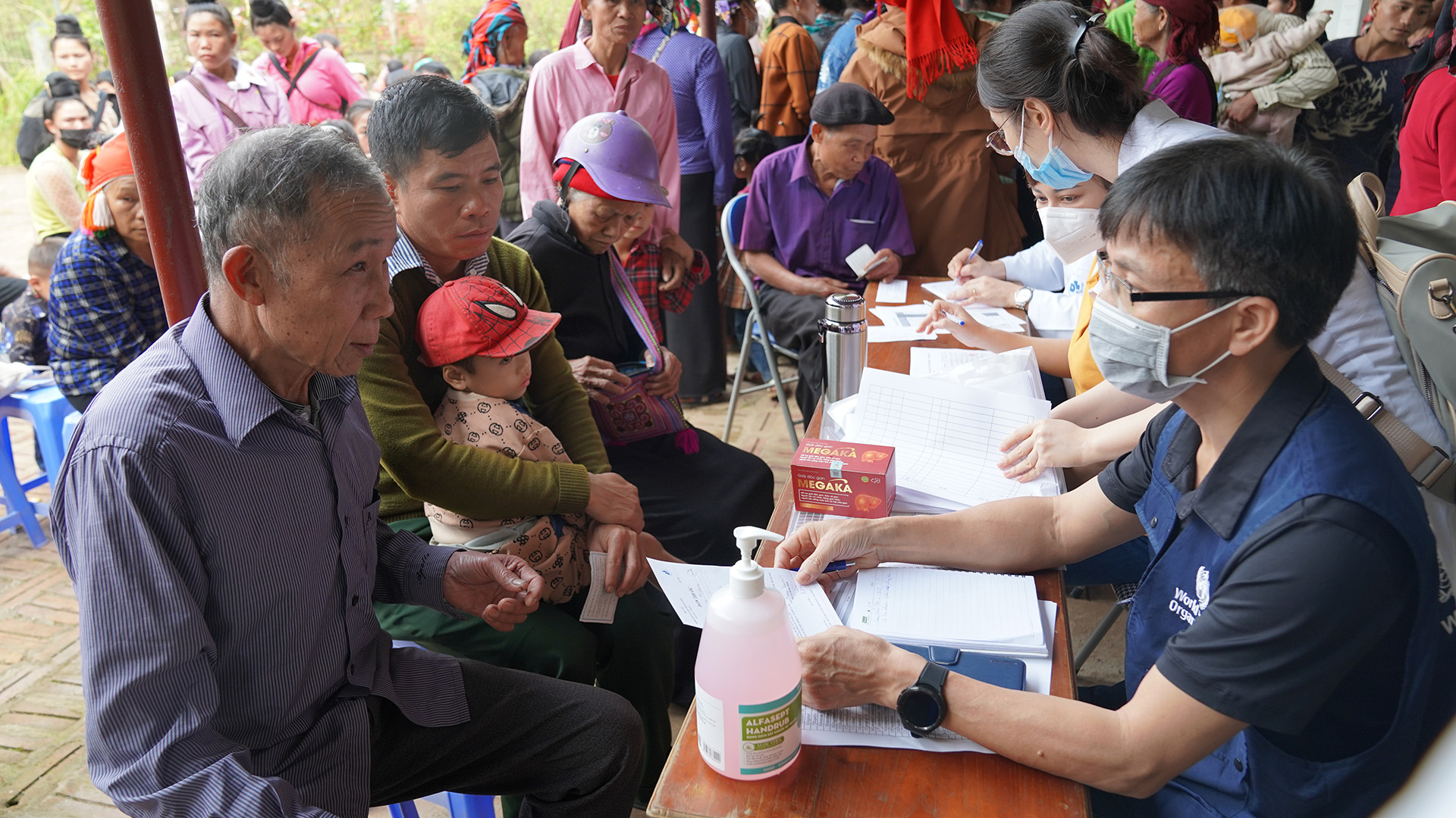 SNY03977 People gathered at a registration table. Individuals at a table are filling out forms. A person holds a child while others wait in line in the background.