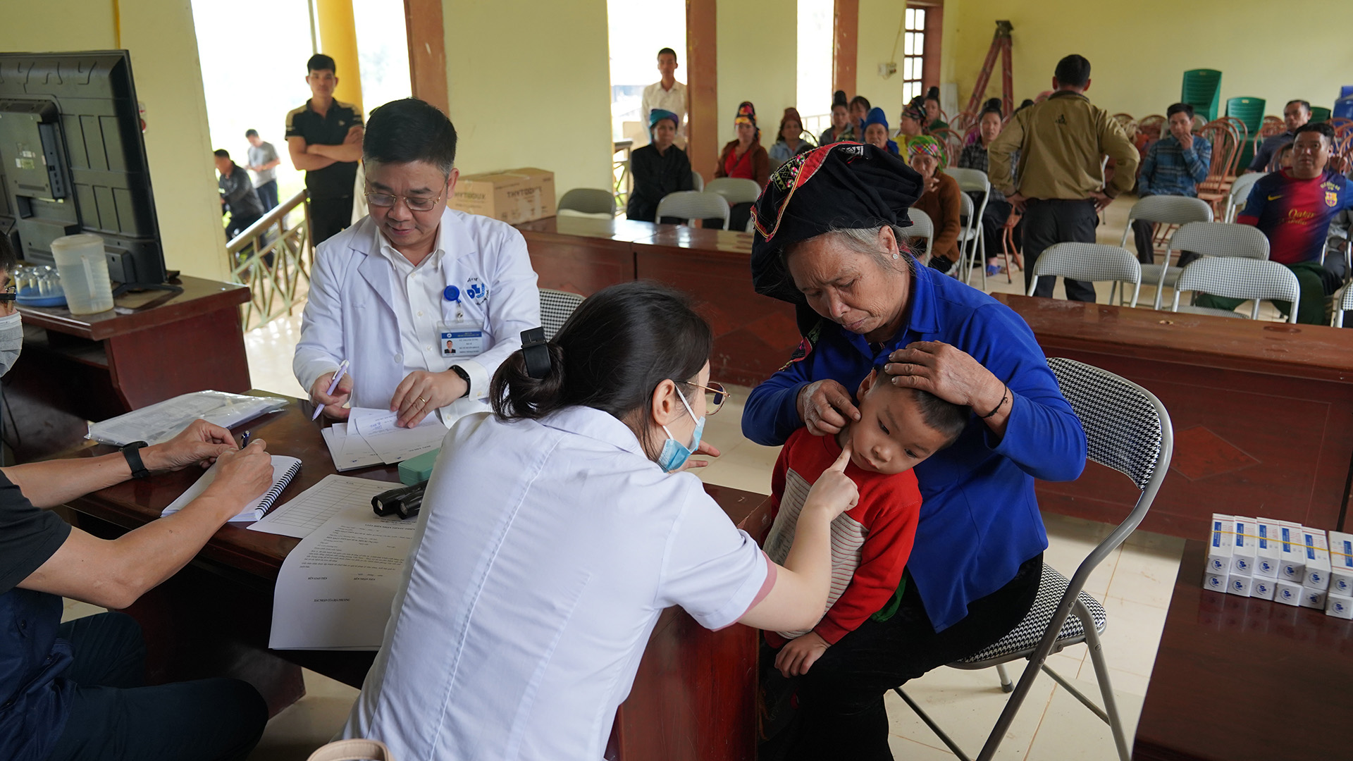 SNY04005 A masked medical professional in a white examines a young child wearing a red and beige sweater, held by an adult in blue attire with a headscarf. Another healthcare worker writes notes at a table, while several people wait seated in the background.