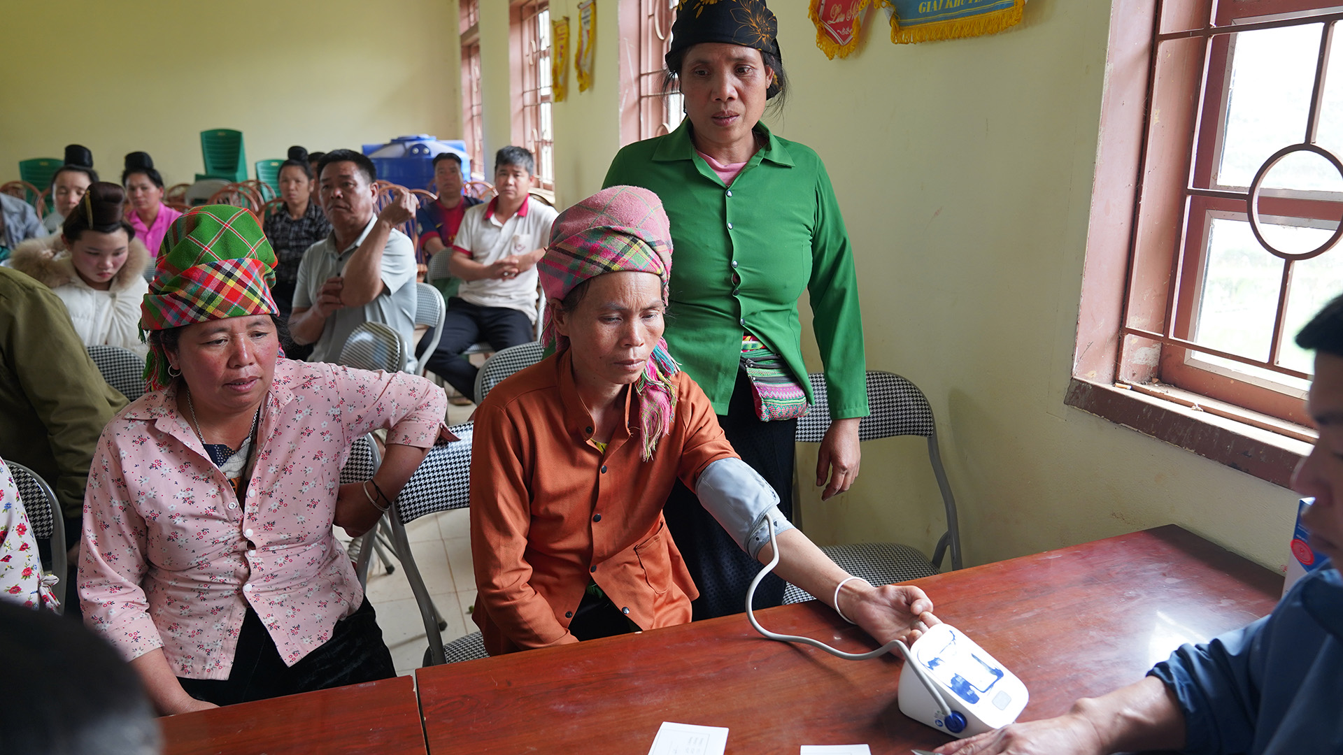 SNY04061 A person in an orange shirt and colourful headscarf has their blood pressure checked using an electronic device. A person in a green shirt stands nearby, while others sit in rows of chairs in the background.
