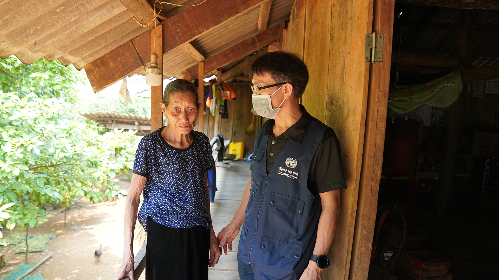 SNY04126 An elderly person in a blue patterned shirt and black trousers stands on the porch of a wooden house, speaking with a World Health Organization worker wearing a dark shirt and WHO vest.