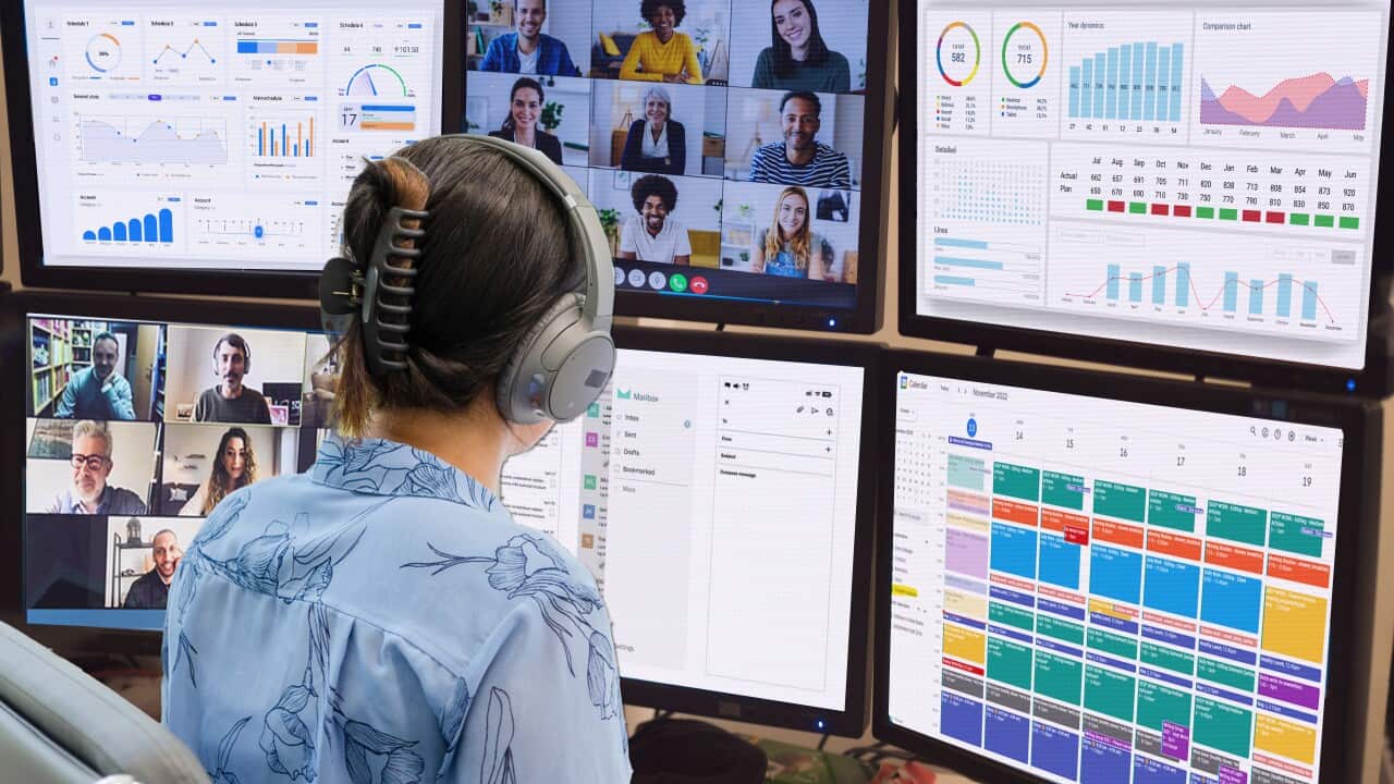 A woman in a blue blouse sits with her back turned, wearing headphones and staring at six computer screens showing graphs, a calendar and two separate video calls