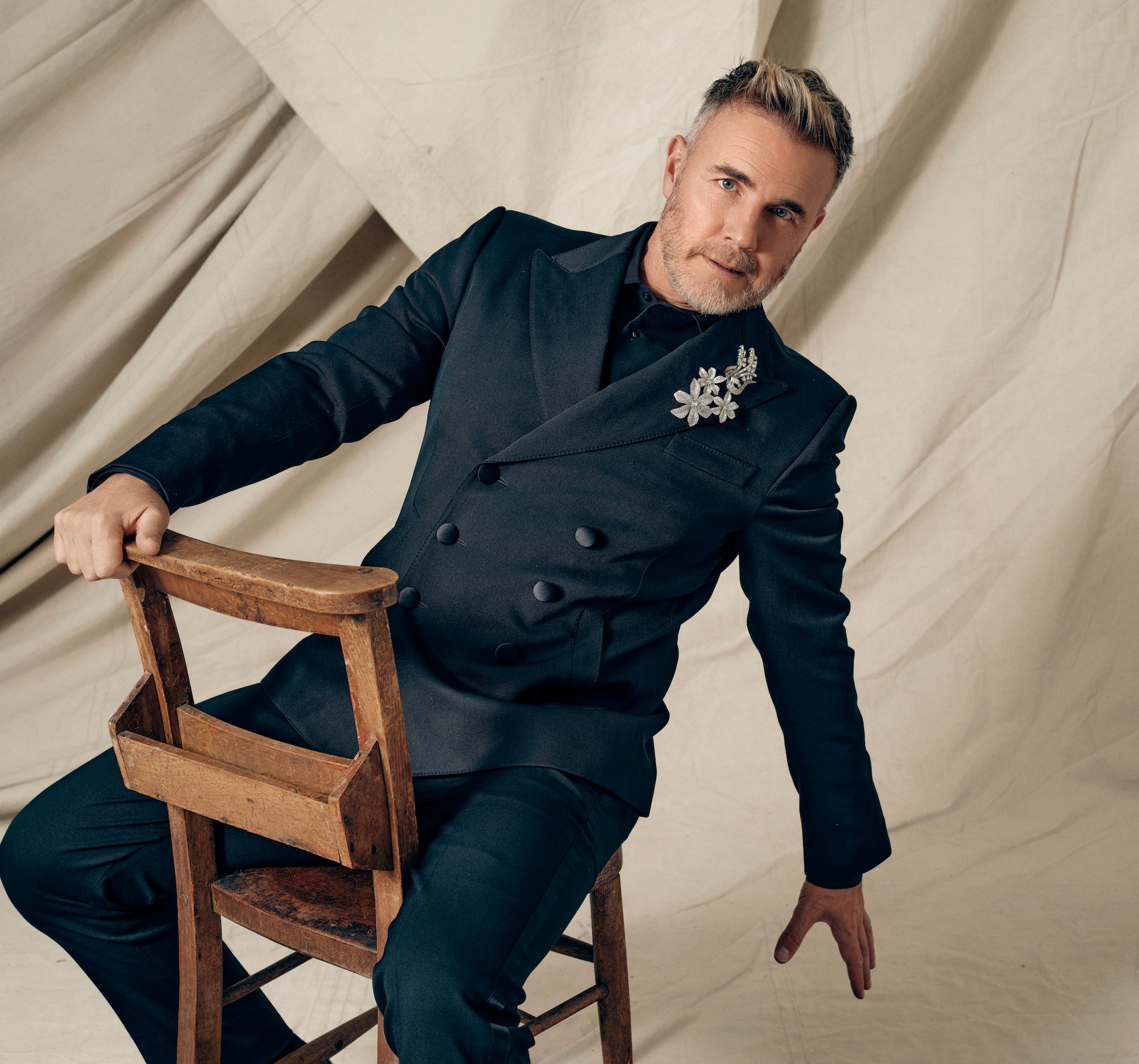 Portrait of Gary Barlow in a black suit sitting on a wooden chair.
