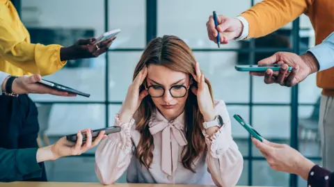 Getty Images Shot of a stressed out businesswoman surrounded by colleagues needing help. Businesswoman feeling stressed out in a demanding office environment at work. Photo of businesswoman under strain as colleagues request various things from her. Businesswoman feeling overwhelmed by her colleagues in the office. Frustrated businesswoman having a headache while her colleagues are demanding her to work on many things in the office. Stress, anxiety and multitasking businesswoman with headache from workload and laptop deadline in office.