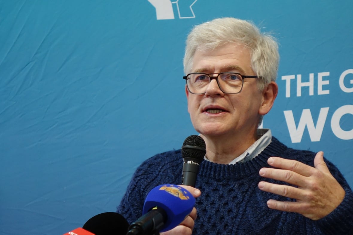 A man wearing a blue knit sweater speaks into a microphone.