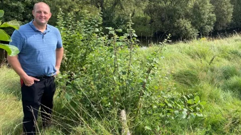 Rob Airey is a stocky farmer in his 30s. He has a blue top and dark jeans and has a shaved head. He is standing with his hands in his pockets next to a tree which has been felled 