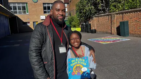 BBC / Gem O'Reilly A father and daughter stood next to each other in a school playground. The father is stood on the left, and has his arm round his daughter. He is looking into the camera. He has dark skin, and is wearing a black coat and black jumper, with a red lanyard round his neck. His head is shaved, and he has a black beard. His daughter is looking at the camera and smiling. She is wearing glasses and has black braided hair. She is holding a copy of the book to the camera, with the title 'My Blood, Your Blood' on the front page, above an illustration of her.