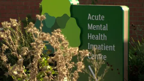 BBC Green signage reading 'Acute Mental Health Inpatient Centre'. The sign is in front of a red brick wall and it is partially covered by a bush. 