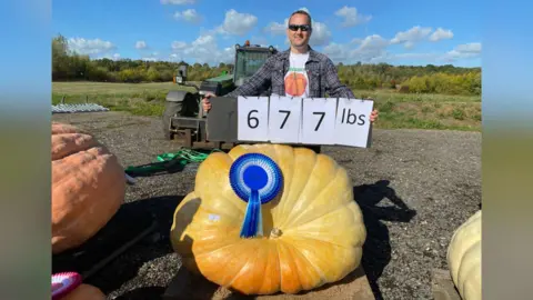 Matt Peskett A man, who is wearing sunglasses, holding up a sign which reads 677 pounds. He is standing next to a giant light orange pumpkin.