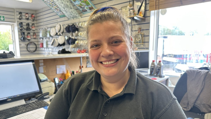 A woman with dark hair tied up smiles at the camera while sat in front of a laptop. She wears a dark green polo-shirt with a motif on a boat on it.