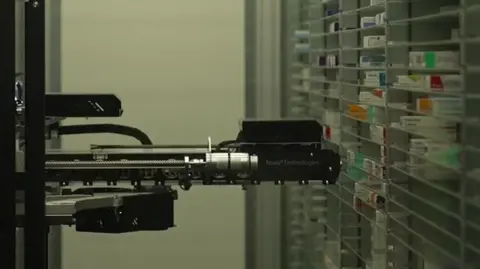 A robotic arm can be seen between two shelves containing boxes of medicine