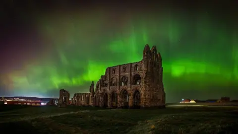 Astro Dog The Northern Lights in green and edged with red over Whitby Abbey in North Yorkshire. The brightest parts look like ribbons in the sky. The ruins of the abbey stand underneath with fields around them. there is a house lit up in the background.
