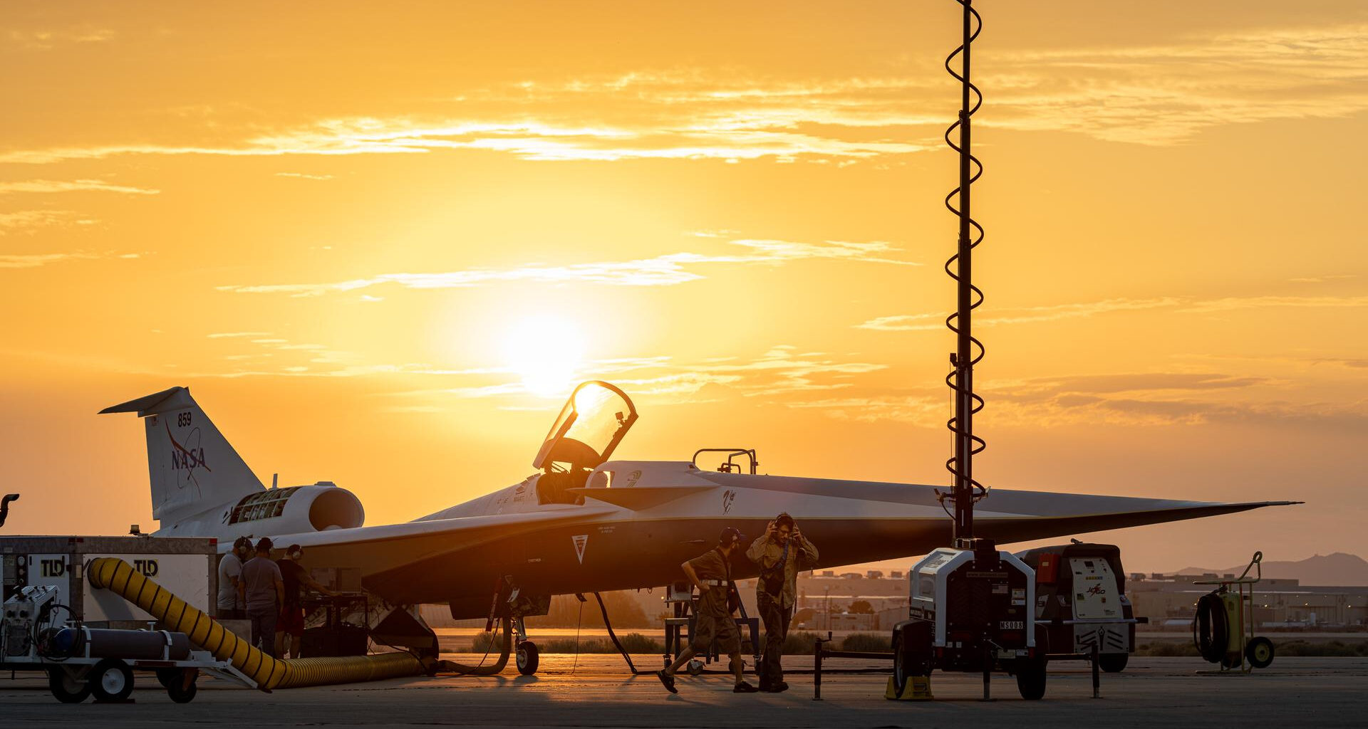 A sleek plane with an open cockpit sits on a landing pad in front of a glowing orange sky with the sunrise.