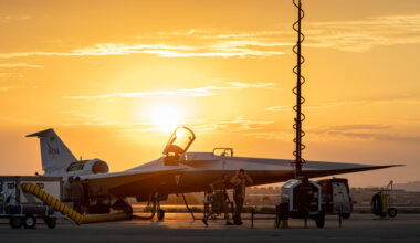 A sleek plane with an open cockpit sits on a landing pad in front of a glowing orange sky with the sunrise.