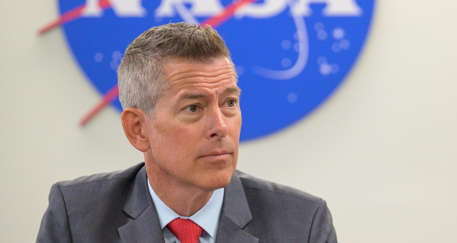 A man in a suit sit looks to the right in front of the bottom half of a nasa meatball logo.