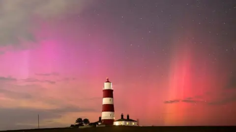 James Rowley-Hill James Rowley-Hill's photograph of pink, red and orange northern lights, shows the aurora over a red and white lighthouse. The cottage next to the lighthouse has its lights on.