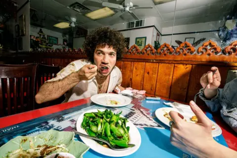 Annie Noelker Benny Blanco sitting in a restaurant putting a spoonful of food into his mouth. A vegetable dish is on the table in front of him.