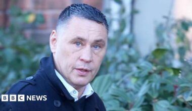 A man glances at a camera as he walks past. He has short cropped hair with gel in it.