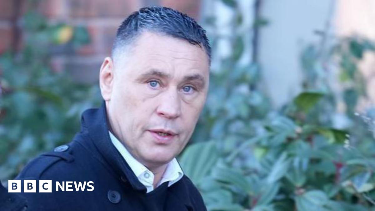 A man glances at a camera as he walks past. He has short cropped hair with gel in it.