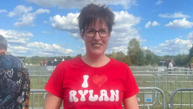 Kelly has short brown hair and is wearing glasses and a red T-shirt that says "I heart Rylan". She is standing in front of rows of small metal barriers.