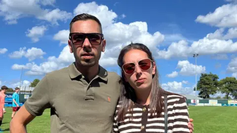 Connor Bennett/BBC Fabio and Rita Serrano on the pitch at Thetford Town FC club. Fabio has short black hair and is wearing dark glasses and a olive green polo shirt. His arm is around his sister who has long black hair and is wearing dark glasses and a black and white stripped cardigan. The sky above is blue with white clouds. 