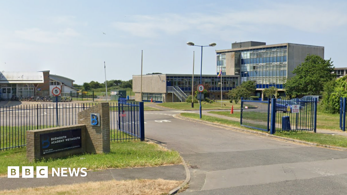 Google Street View of Budmouth Academy in Weymouth. Blue metal gates open onto a drive surrounded by buildings. The largest is a late 20th-century four-storey block covered in glass and blue panels and with a flat roof.