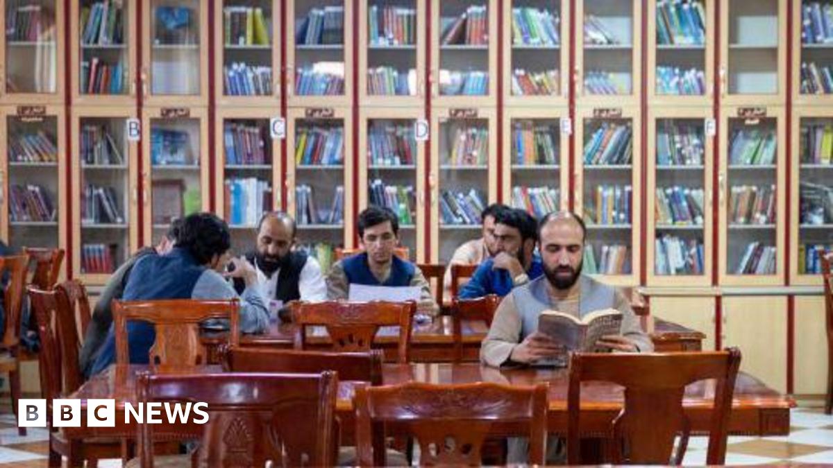 Men sit at tables in a room with bookshelves in the background