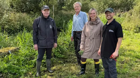 Michael Barrett Four people, standing outside, on grass, by vegetation, all smiling and looking at the camera. There are three men and one woman. 