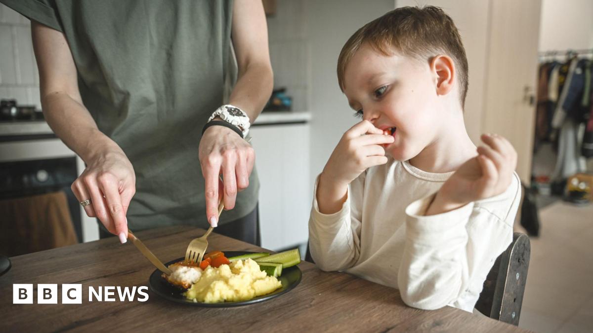 A young child with brown hair and a white top, eating a tomato, with a woman wearing a green top cutting up a plate of food in front of him.