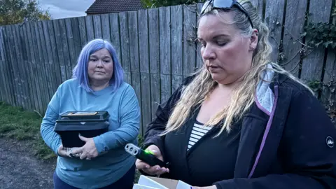 Jim Scott/BBC Julie Kilmartin, left, and wearing a blue top is holding a box which houses Nancy while Lauren Davison, right, is holding a bat detector which senses when similar breeds are nearby.