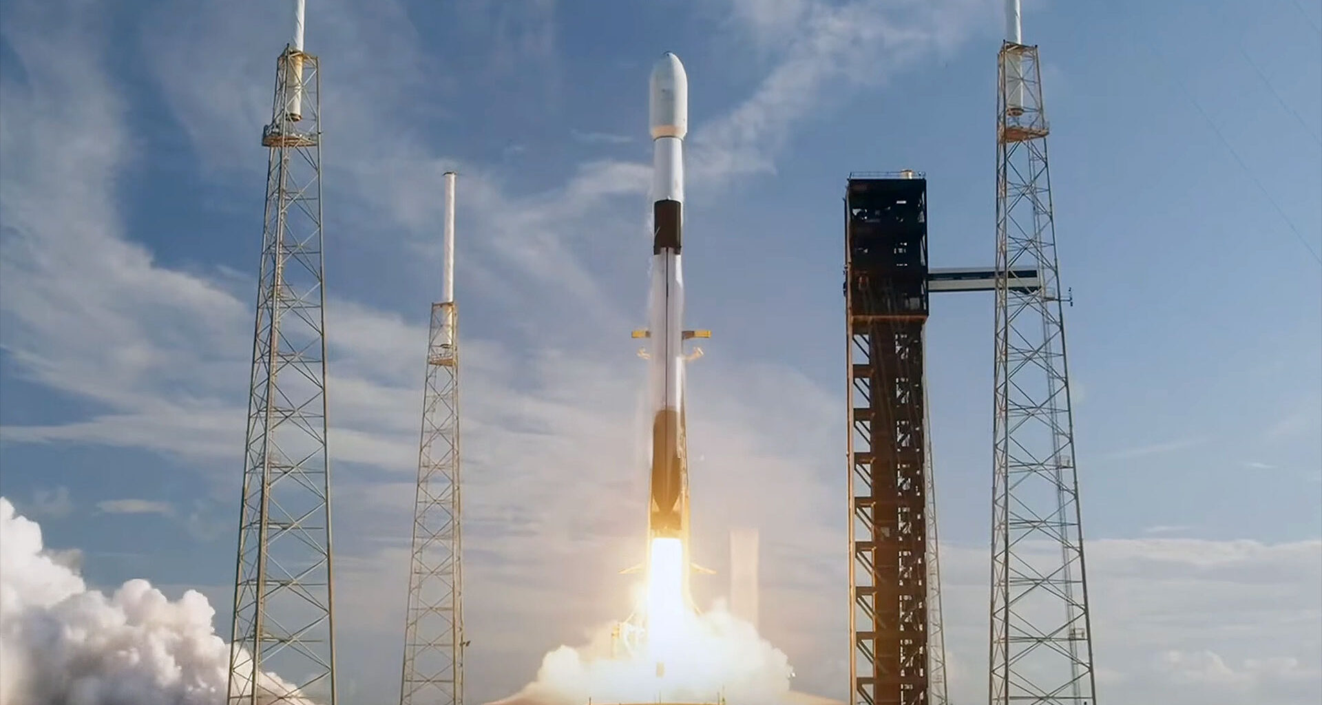 a white and black rocket lifts off on a bright plume with an orange glow, set against a blue sky with wispy white clouds.