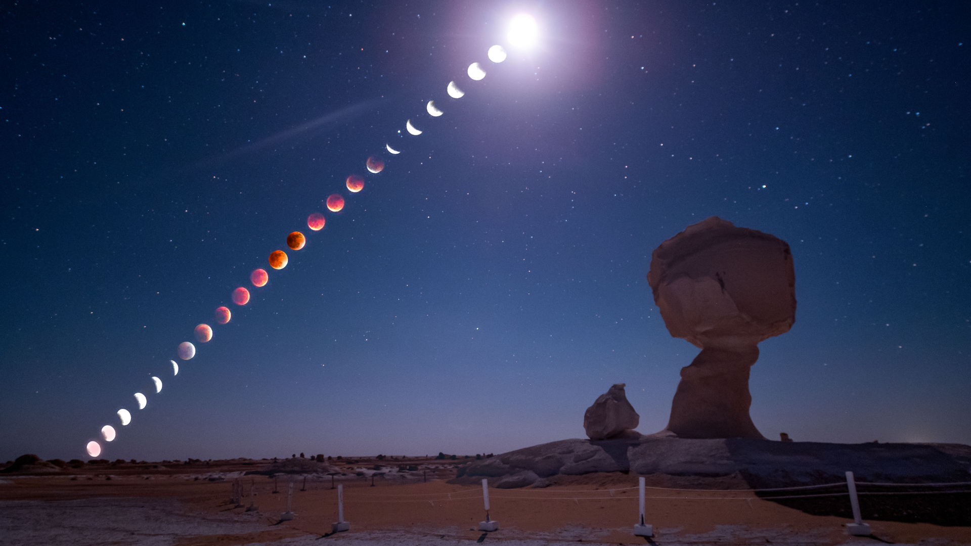 A composite image showing the phases of a blood moon total lunar eclipse unfolding in a line through a starry sky above a desert featuring two large chalk formations.