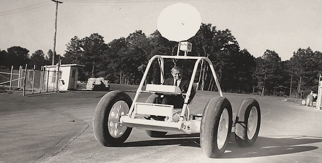 A black and white photo showing a man in a suit in a four-wheeled car rolling down a road.