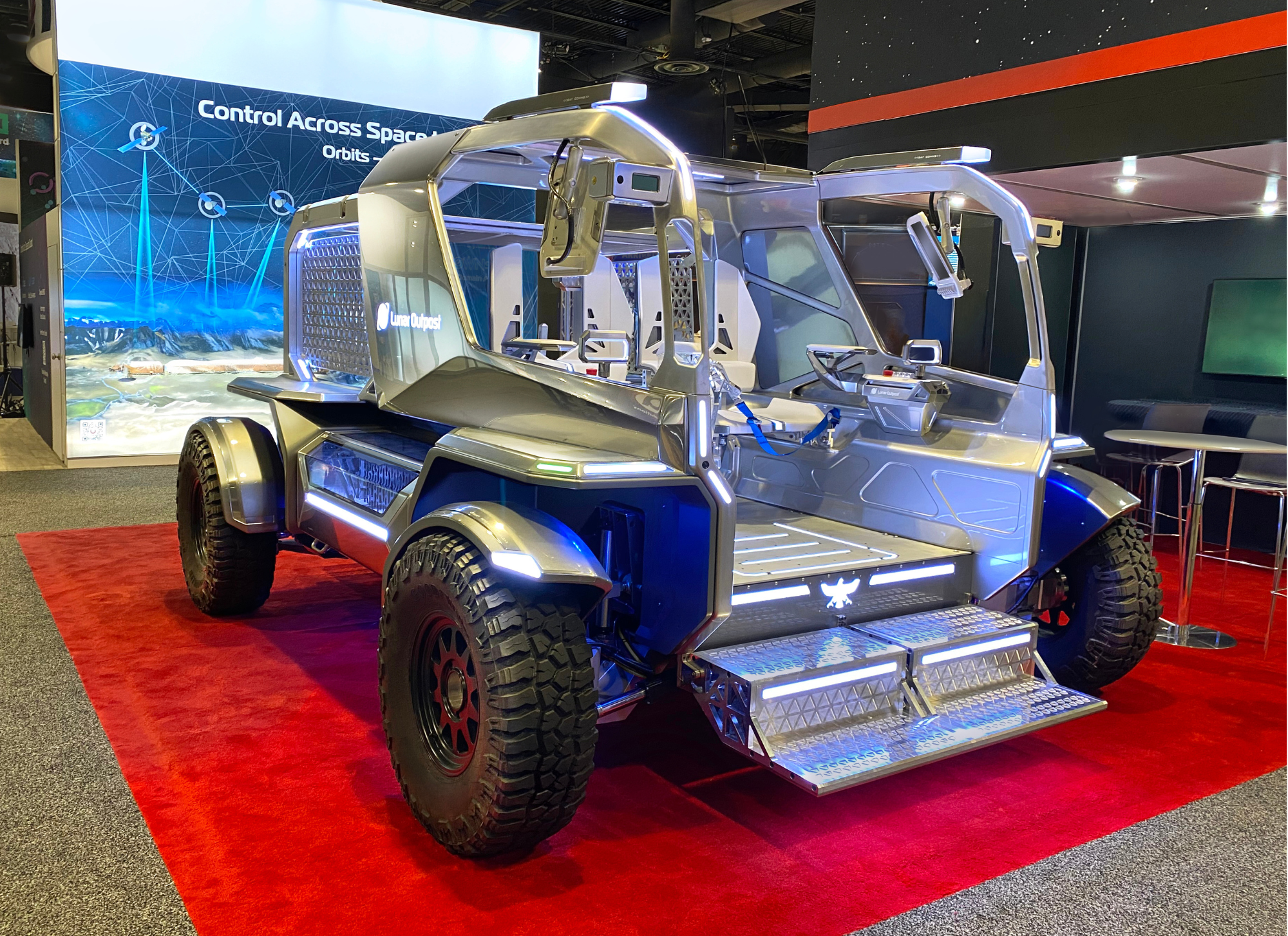 A large four-wheeled rover made of metal with four seats sits on a square of red carpet with a background and display lights