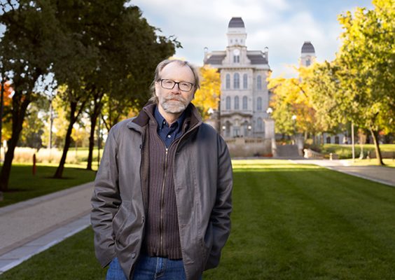 George Saunders, man in grey jacket standing on campus with Hall of Languages and fall foliage in the background
