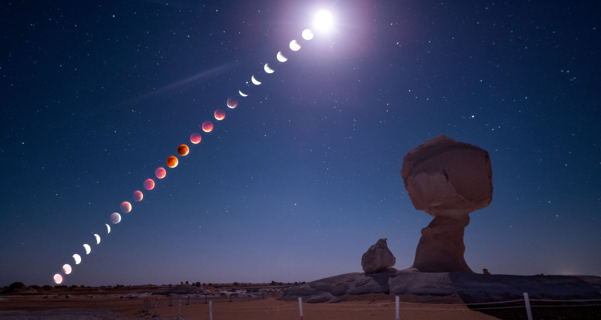 A composite image showing the phases of a blood moon total lunar eclipse unfolding in a line through a starry sky above a desert featuring two large chalk formations.