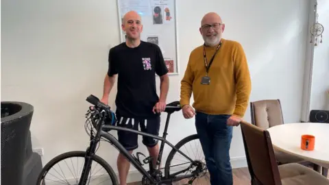 Men Only IOW (l-r) Dave Walters wearing black cycling shorts and a black t-shirt is holding onto his bike. He is standing next to Karl Hart who is wearing a yellow jumper and blue jeans at the open space centre in Ryde.