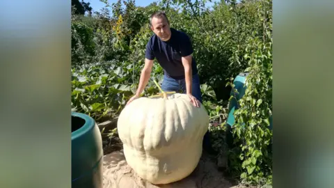 Matt Peskett A man in a blue shirt and jeans standing over a giant white pumpkin. He is standing inside a garden.