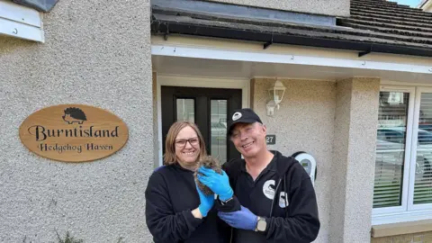 Sharon Longhurst Sharon holds a hedgehog. She is wearing blue rubber gloves. Her husband Andy is standing beside her and smiling. He is wearing a black baseball cap and a dark top. She is wearing a dark jumper and is also smiling. They are standing in front of their house which has a sign on it that says Burntisland Hedgehog Haven.
