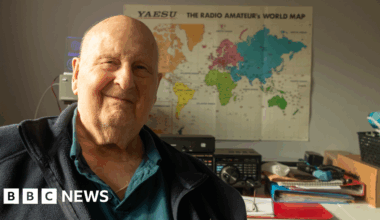 92-year-old Ray Pratt, sitting smiling in front of his home radio setup.