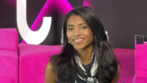 Anja-Sara Lahady Smiling, with long dark hair Anja-Sara Lahady stands in front of pink cushions.