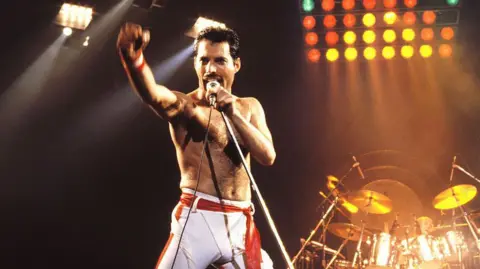 Getty Images Freddie Mercury sings on stage with his top off and holds his arm in the air 