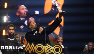 Scorcher stands on stage holding a microphone in one hand and an award in the other. He is holding the award in the air. There are four other men on the stage behind him. Two of them are filming him on their mobile phones.