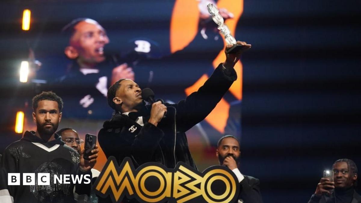 Scorcher stands on stage holding a microphone in one hand and an award in the other. He is holding the award in the air. There are four other men on the stage behind him. Two of them are filming him on their mobile phones.