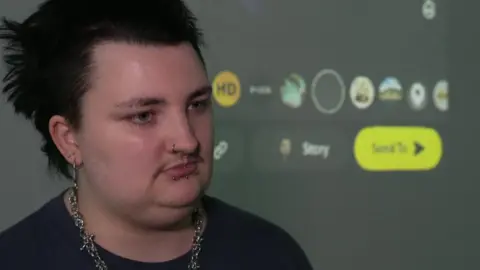 Arron is talking whilst standing in front of a projector screen. He is wearing a black t shirt.